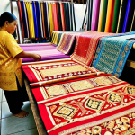 **A vibrant, detailed photograph of Songket fabrics displayed at a Brunei crafts market. The gold and silver threads should shimmer, showcasing the intricate patterns. Focus on the rich textures and the traditional weaving techniques. In the background, blurred figures of local artisans can be seen working on looms. High resolution, natural lighting.**